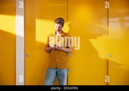Male business entrepreneur using smart phone while standing near yellow wall in office Stock Photo