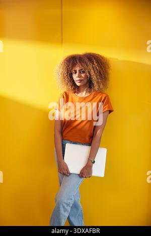 Portrait of confident female business expert holding laptop while standing near yellow wall Stock Photo