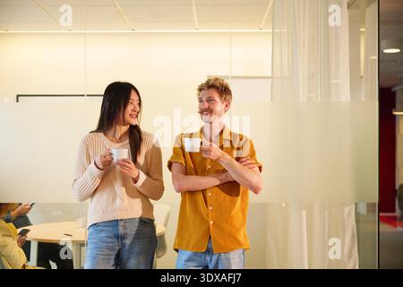 Smiling male and female business experts having coffee while talking with each other standing in office Stock Photo