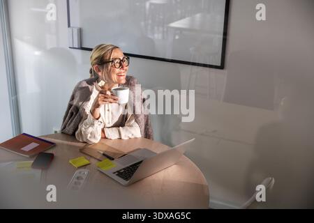 Happy businesswoman holding coffee cup and looking up while sitting at office Stock Photo
