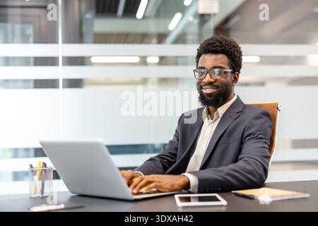 Young professional man wearing eyeglasses typing on a laptop, looking at camera and smiling while sitting at his desk in a contemporary corporate environment Stock Photo