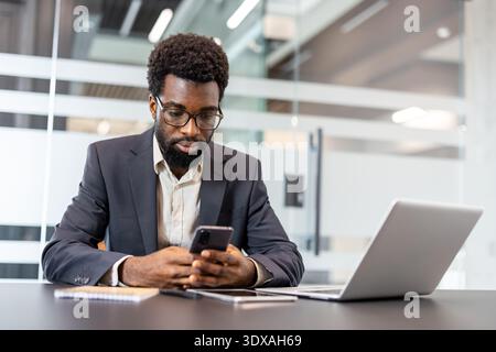 Young businessman engaged with cellphone technology at a modern desk, multitasking between digital devices while working in a bright corporate environment, symbolizing connectivity and productivity Stock Photo