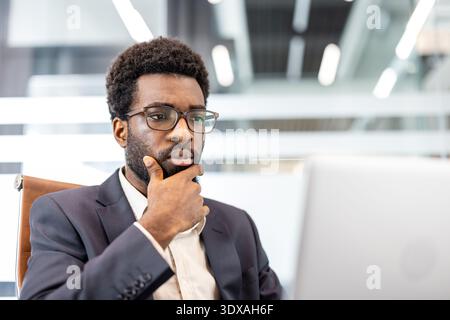 Thoughtful businessman focusing on a silver laptop screen, developing new ideas and concepts while working in a modern office environment, fostering innovation and success Stock Photo