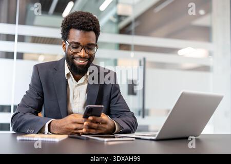 African man with glasses smiling, holding and using a smartphone while sitting at a modern office desk with a laptop and tablet, engaged in technology and communication Stock Photo