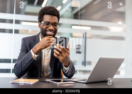 Smiling businessman browsing smartphone at a desk with open laptop in a modern office, looking positive and connected while working remotely and staying productive and professional Stock Photo