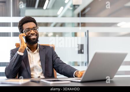 Smiling professional man talking on cell phone while working on laptop in a modern office, engaged in clear communication and successful business dealings Stock Photo