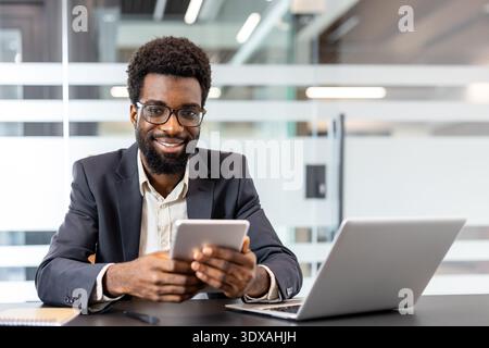 Cheerful male professional looking at camera and holding digital tablet while sitting at desk in an open-plan contemporary workspace, representing success, management, and global business technology Stock Photo