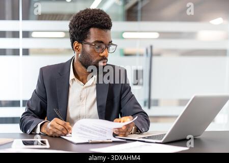 Concentrated professional man reviewing documents and working on a laptop at a modern office desk, managing business affairs and planning new strategies Stock Photo