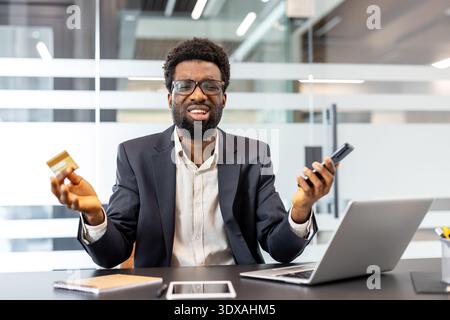 Businessman sitting at a desk in an office, holding a credit card and smartphone while looking overwhelmed by a payment issue or financial problem, indicating debt or bankruptcy Stock Photo