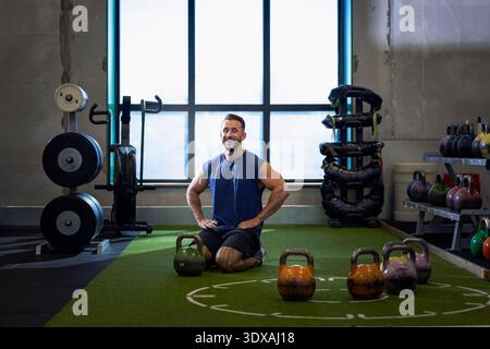 Smiling man with arms akimbo kneeling on floor while working out in gym Stock Photo