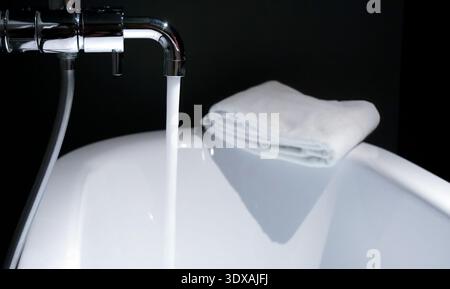 A clean folded white towel rests on the rim of a white ceramic bathtub while water flows from a chrome faucet. Set against a dark background, this ima Stock Photo