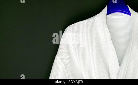 Clean white cotton bathrobe with waffle weave texture hanging on a blue hanger against a black wall. Minimalist composition with ample negative space Stock Photo