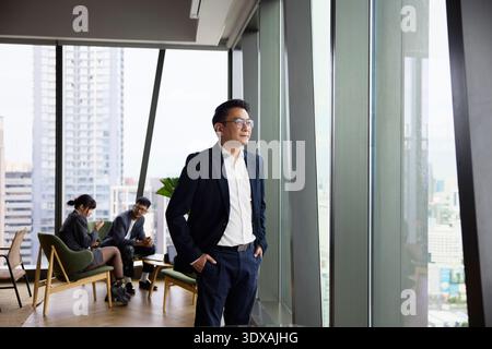 Confident businessman looking through window while standing with hands in pockets at office Stock Photo