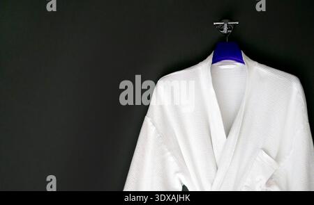 Clean white cotton bathrobe with waffle weave texture hanging on a blue hanger against a black wall. Minimalist composition with ample negative space Stock Photo