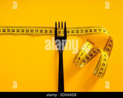 Black fork and yellow measuring tape on a yellow background, symbolizing healthy eating, portion control, and fitness goals Stock Photo