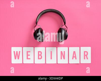 Black wireless headphones on pink background with WEBINAR spelled out on white cards. Listening to webinar and online learning concept. Stock Photo