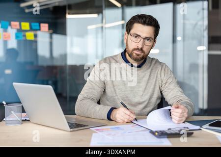 Businessman checking documents and statistics, wearing glasses and a beard while working at his desk with a laptop and calculator in a contemporary glass office Stock Photo