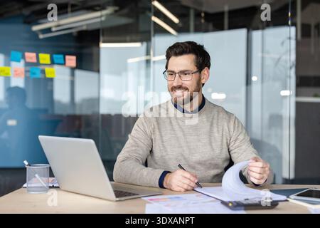 Smiling man in eyeglasses sitting at a desk, looking at his laptop screen while holding a pen and flipping through papers, focusing on his work in a modern office Stock Photo
