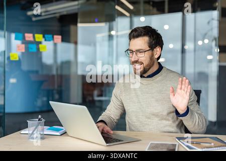 Smiling businessman wearing glasses communicating with colleagues on a laptop, working at a desk in a modern office, using video conferencing for remote connection Stock Photo