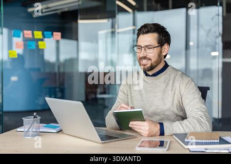 Happy male professional working from modern office, collaborating on a project, writing information in a notebook while looking at a laptop computer screen during an online meeting Stock Photo