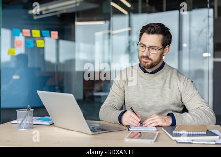 Businessman wearing glasses diligently working on a laptop computer and taking notes, in a notebook at his desk in a contemporary co-working office space with glass walls Stock Photo