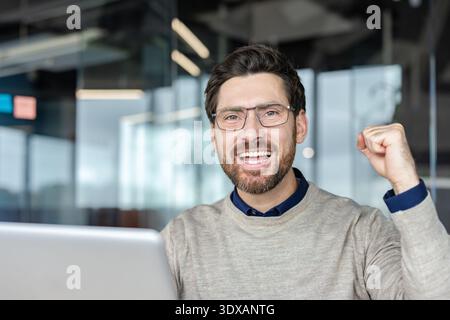Confident businessman celebrating achievement, showing joy and excitement, making a fist pump gesture while working on a laptop in a modern office environment Stock Photo