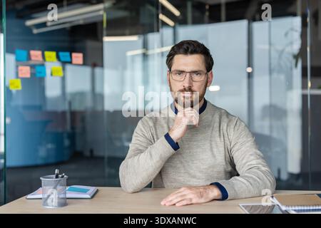 Serious professional businessman actively thinking and planning, sitting at a modern office desk with a thoughtful expression and hand on chin, representing business intelligence and problem-solving Stock Photo