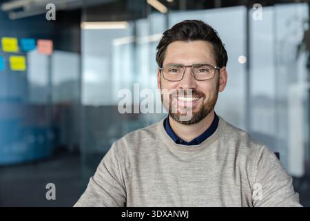 Smiling businessman wearing casual sweater and modern glasses posing for a portrait in a contemporary office, representing corporate success, confidence, and professionalism Stock Photo