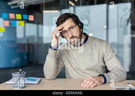 Frustrated call center worker wearing a headset and glasses, holding his head, experiencing stress and headache while looking at the camera in a modern office setup Stock Photo
