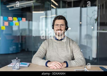 Smiling male call center agent wearing a headset and glasses, sitting at his desk in a modern office, providing distant customer service and contact support Stock Photo