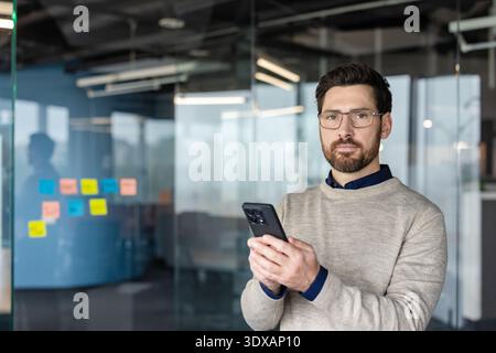 Confident businessman standing in a modern glass office, wearing eyeglasses and a sweater, looking at the camera while holding his smartphone and communicating Stock Photo