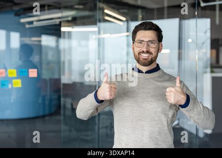 Smiling man in an office setting giving two thumbs up, expressing success, approval, positive feedback, and agreement in a professional business environment Stock Photo