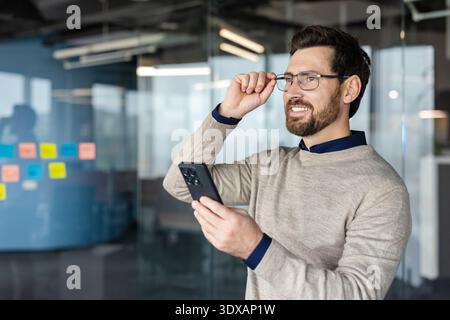 Young businessman holding a mobile phone and adjusting his spectacles while smiling and looking away in a contemporary office environment, representing business vision and technology use Stock Photo