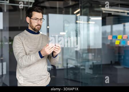 Young businessman wearing eyeglasses and a casual sweater concentrating on a digital tablet, working and managing information in a contemporary corporate office environment Stock Photo