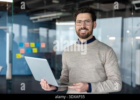 Smiling professional businessman wearing glasses and casual sweater standing confidently and looking at camera, holding a laptop in a contemporary office workspace Stock Photo
