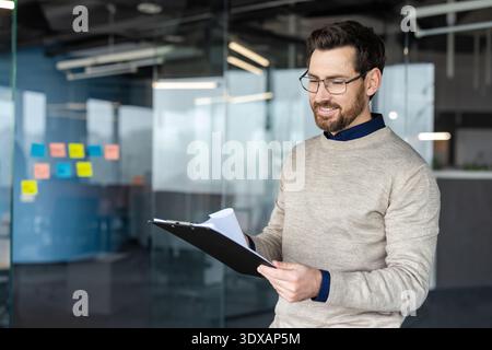 Young businessman smiling while carefully examining documents on a clipboard, standing in a modern office environment with glass walls and sticky notes Stock Photo