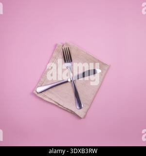 A fork and knife are placed on a beige napkin against a pink background. The utensils are stainless steel with a shiny finish. Stock Photo