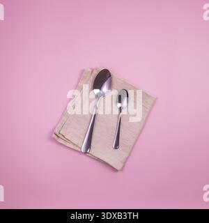 A set of silver spoons placed on a beige napkin against a pink background. The spoons vary in size, showcasing a simple and elegant dining setup. Stock Photo