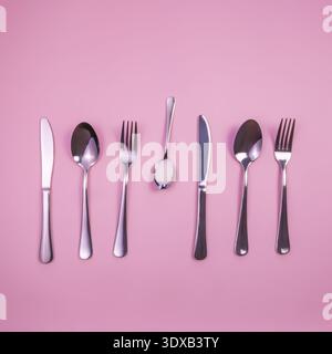A collection of stainless steel cutlery arranged on a pink background. Includes knives, forks, and spoons of various sizes and shapes. Stock Photo