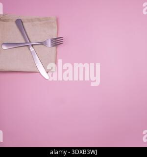 A silver knife and fork are crossed on a beige napkin, placed on a pink background. The scene is simple and minimalistic, focusing on dining utensils. Stock Photo