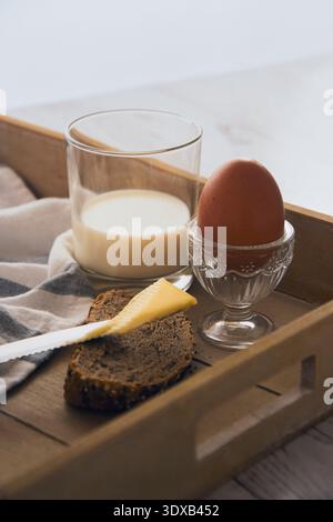 Simple breakfast with a glass of milk, a soft-boiled egg in a decorative glass egg cup and a slice of bread with a knife spreading butter on a wooden Stock Photo