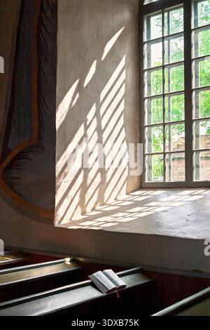 Church interior with sunlight streaming through a window casting shadows on wooden pews with an open bible Stock Photo
