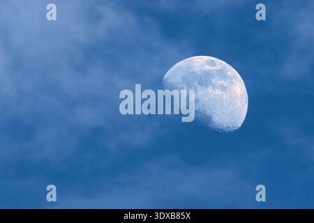 Close-up of a gibbous moon visible during the day, partially covered by soft wispy clouds in a blue sky. Stock Photo