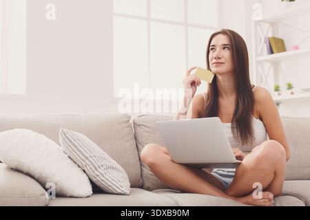 Young Woman Sits Comfortably on a Couch With a Laptop and a Note While Deep in Thought Stock Photo