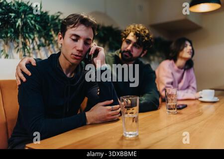 Three friends in a cafe at a wooden table, one on the phone, others seated nearby Stock Photo