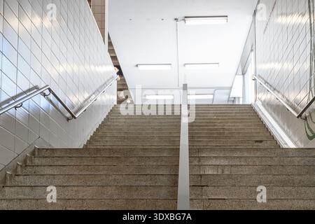 A concrete staircase with metal handrails, tiled walls, ceiling lights, and corridor interior. Stock Photo