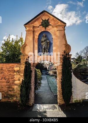 Ornate terracotta archway with Virgin Mary statue in blue niche topped by star motif flanked by cypress trees leading into Portmeirion gardens under s Stock Photo