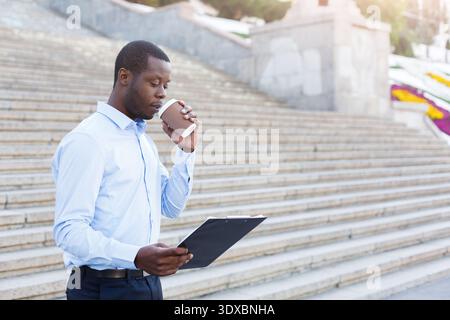 Man Holding a Clipboard and Coffee Stands on Steps in an Outdoor Setting During the Day Stock Photo