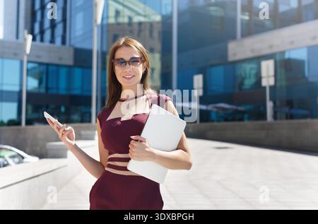 Young Woman Stands Outside Modern Building, Using Phone and Holding Laptop Stock Photo