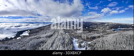 Aerial view of a snow-laden forest stretches across rolling hills towards a distant lake under a sky of mixed clouds and bright blue, Soboth, Styria, Stock Photo